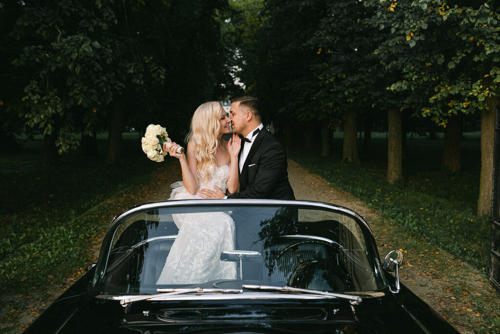 A bride and groom sit in a convertible on a tree-lined path. The bride holds a bouquet of white flowers, and both are dressed in formal attire, smiling at each other.