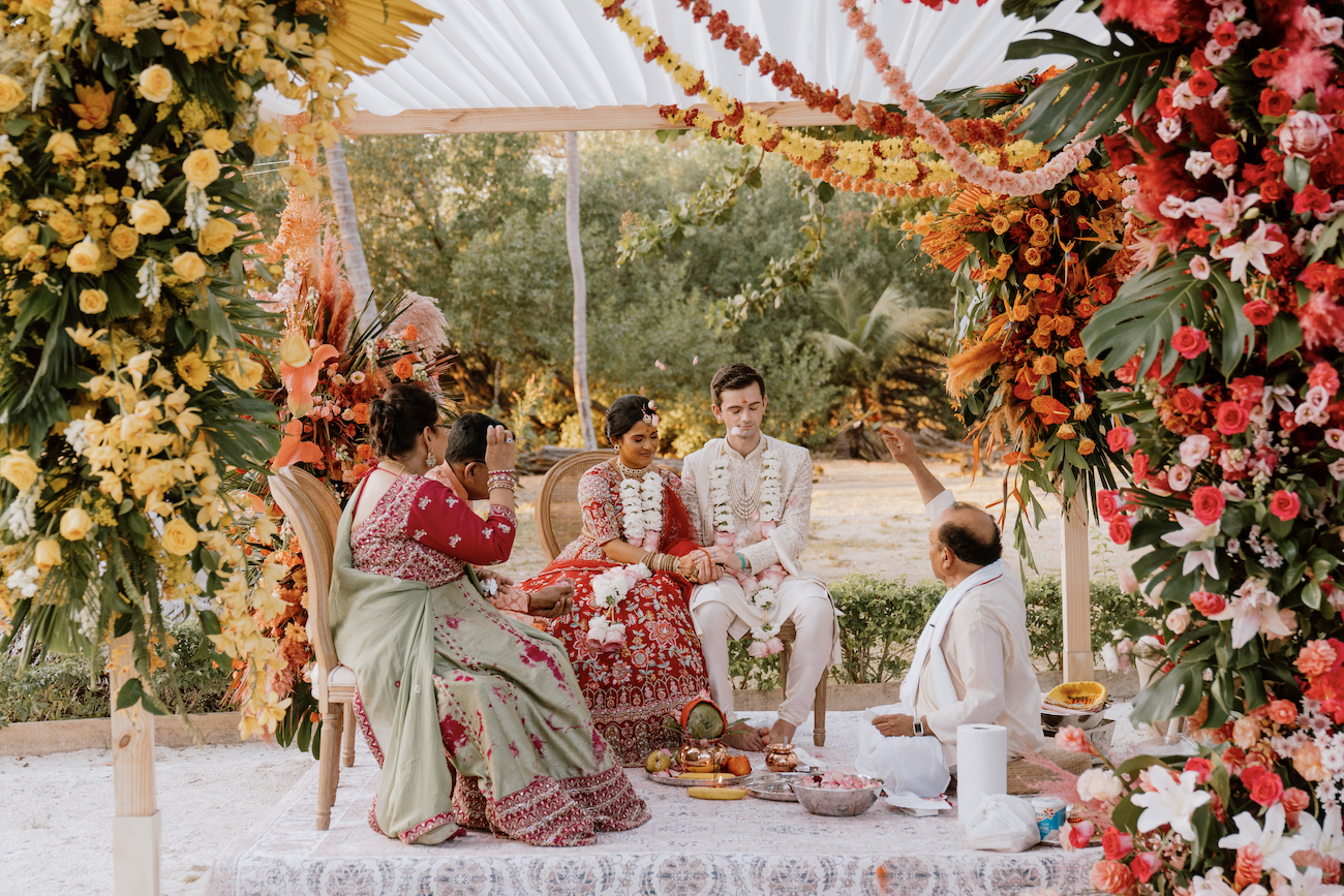 A couple sits together in traditional attire during an outdoor Indian wedding ceremony, surrounded by colorful floral decorations and family members.