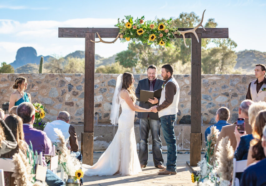 A bride and groom stand under a decorated wooden arch with sunflowers during an outdoor wedding ceremony, with guests seated on either side.