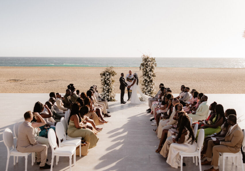 A beach wedding ceremony with a couple standing under floral arches. Guests are seated on either side of the aisle, facing the ocean.