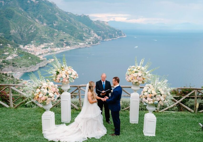 Outdoor wedding ceremony overlooking a coastal landscape. A couple stands before an officiant, surrounded by flower arrangements.