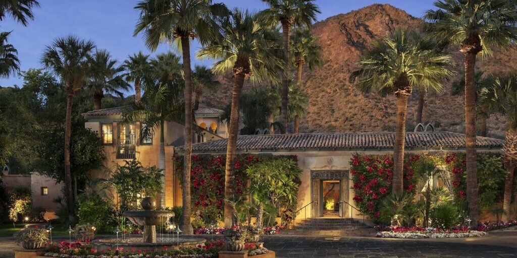 A Mediterranean-style villa with palm trees, flowering vines, and a courtyard fountain sits at the base of a rocky mountain at dusk.
