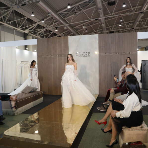 Three models walk on a runway wearing elegant white gowns in a well-lit fashion show setting with an audience seated nearby.