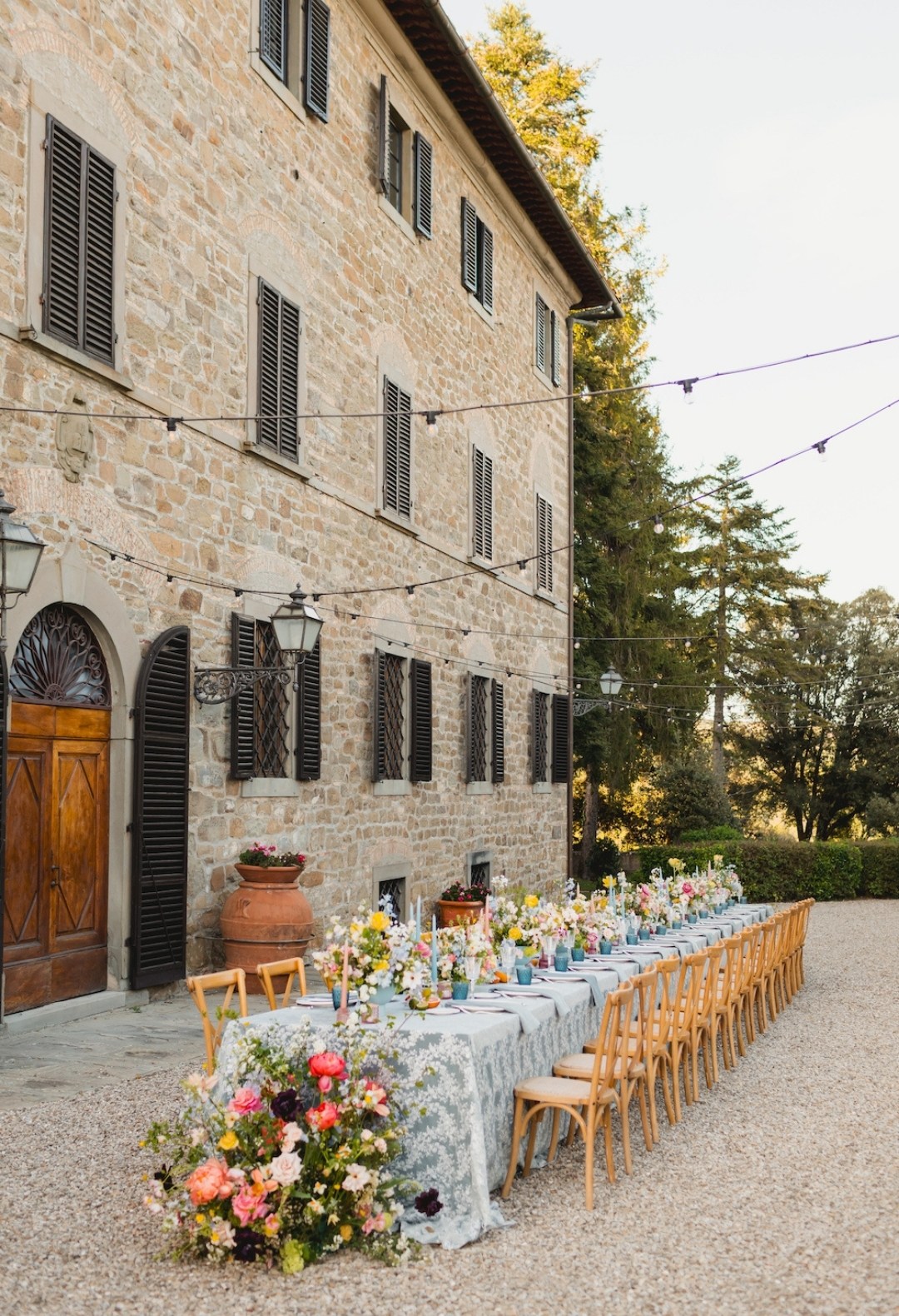 A long outdoor dining table with floral decorations and wooden chairs is set beside a stone building under string lights.