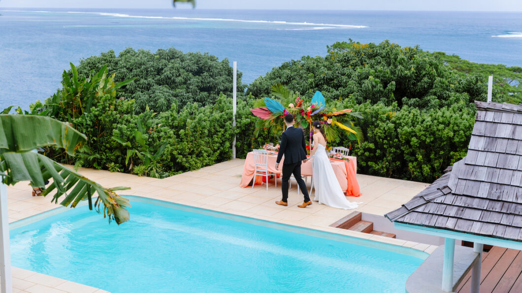 A couple stands by a decorated table near a pool overlooking the ocean in Tahiti, surrounded by lush greenery.