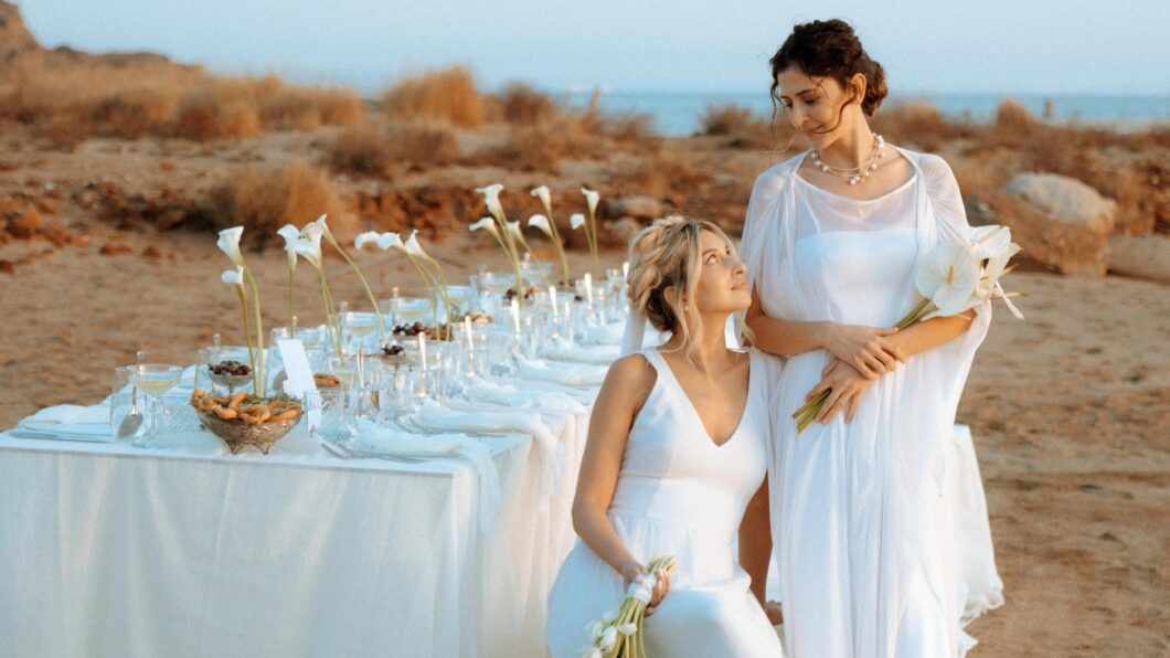 Two women in white dresses pose beside an outdoor table set with white flowers and tableware on a beach, with dry grass and the sea in the background.