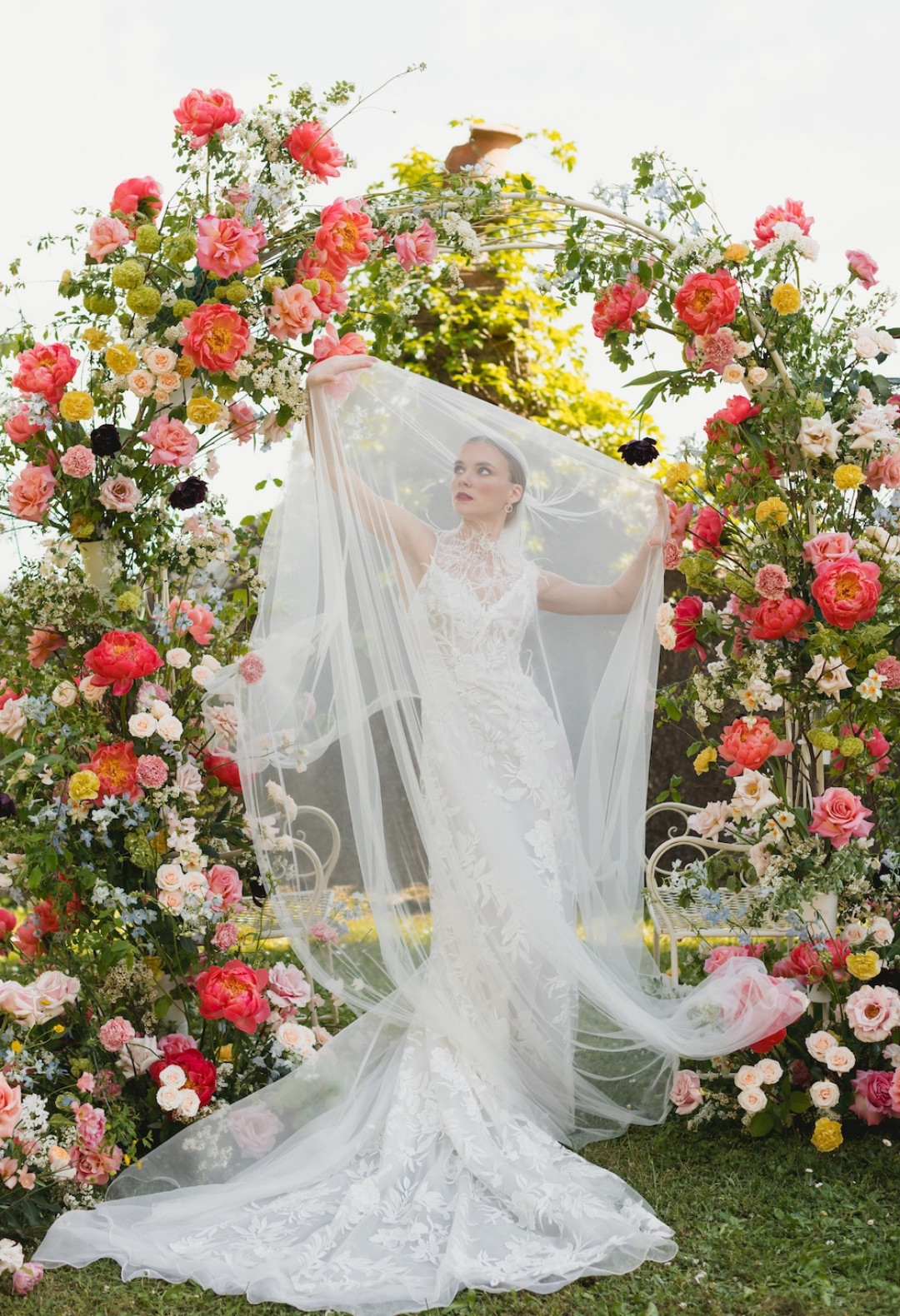 A bride in a lace wedding gown holds up her veil, standing under a floral arch with vibrant pink, red, and yellow flowers in an outdoor setting.