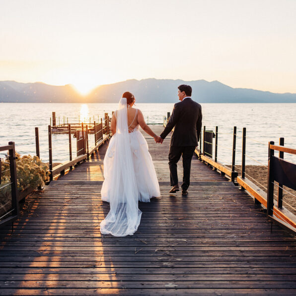 A bride and groom walk hand in hand on a wooden pier at sunset, with mountains and a lake in the background.
