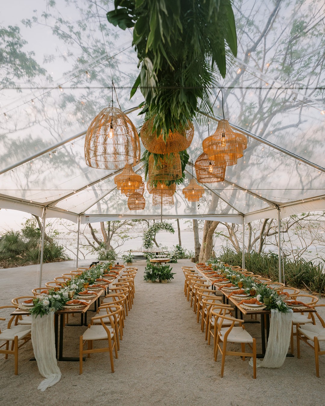 Open-air wedding ceremony setup under a clear tent with wooden chairs, long tables, greenery, and woven pendant lights, overlooking a natural outdoor setting.