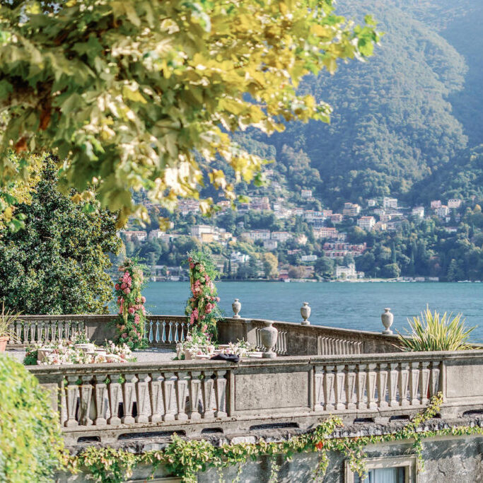 A stone terrace with floral decorations overlooks a lake and distant hillside town, surrounded by greenery under a partly shaded sky.