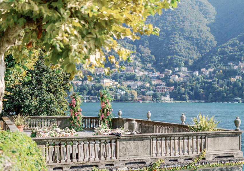A stone terrace with floral decorations overlooks a lake and distant hillside town, surrounded by greenery under a partly shaded sky.