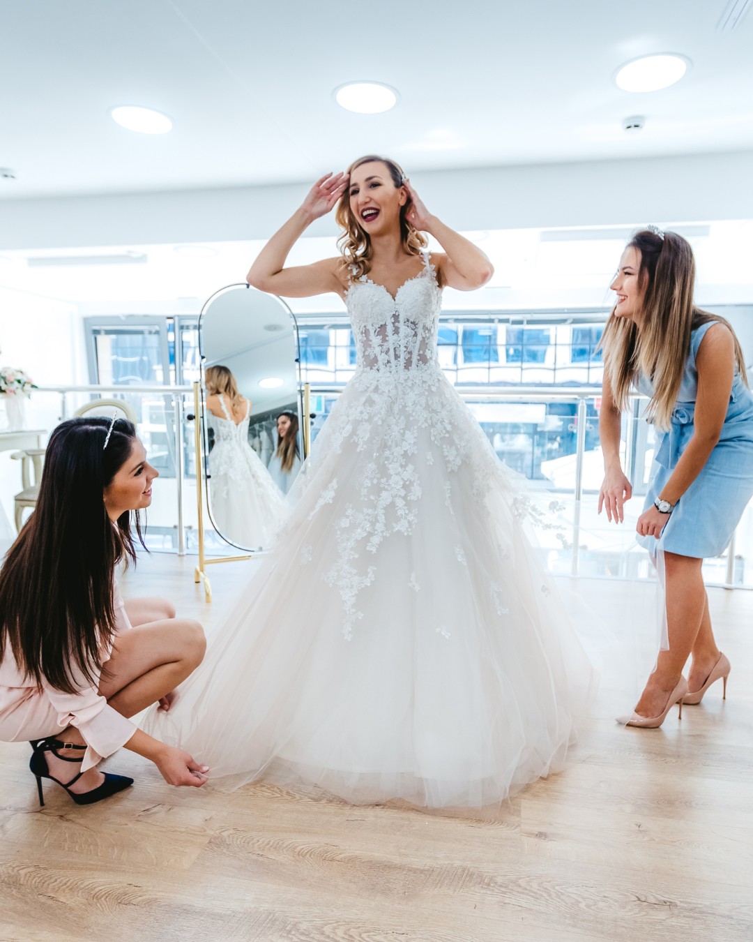 A bride smiles while trying on a lace wedding dress. Two women assist her by adjusting the gown at a bridal shop.