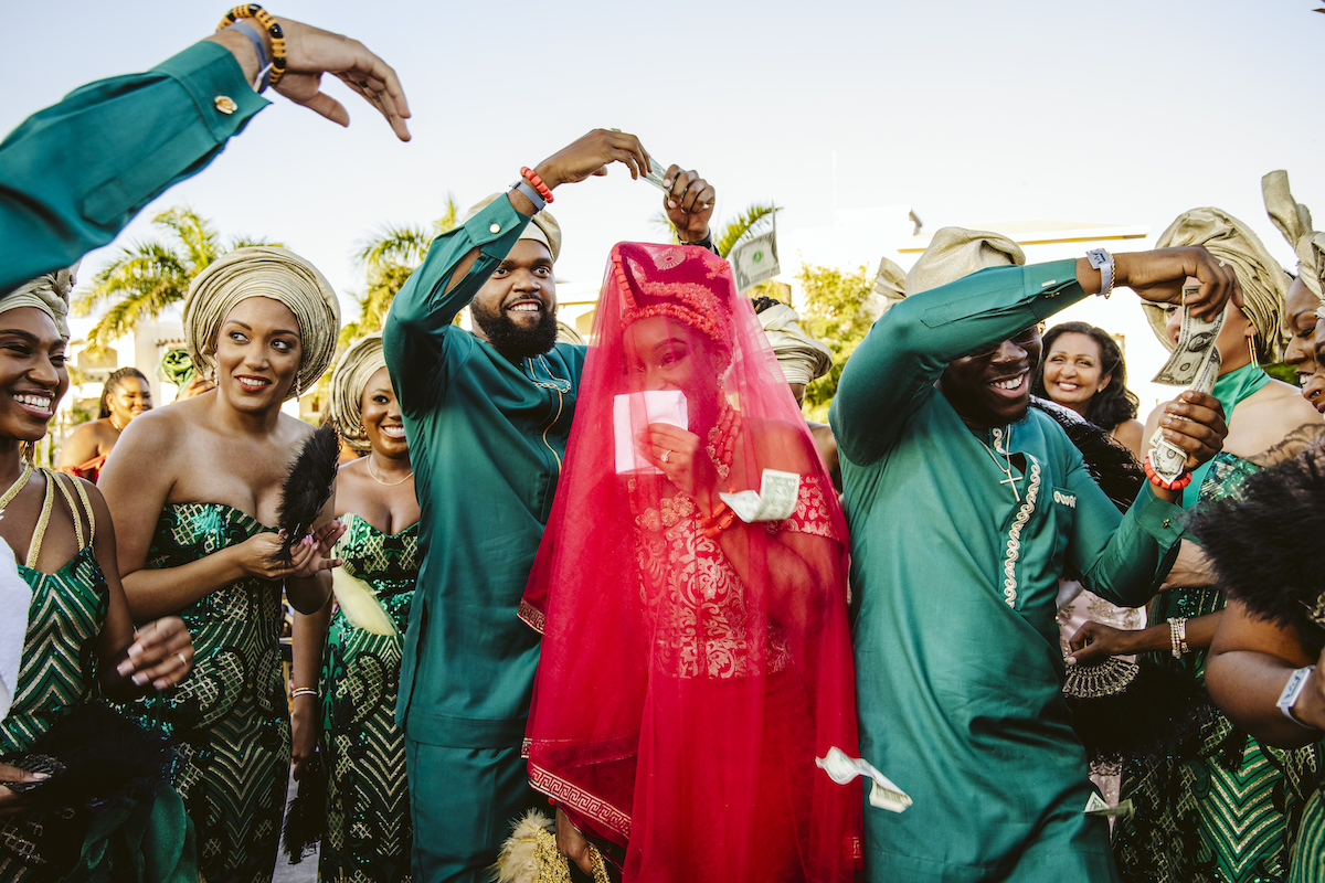 A bride covered with a bright pink veil is surrounded by people in green traditional attire during a festive outdoor celebration.