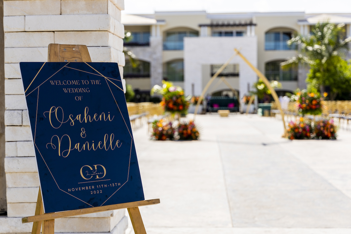 A decorative wedding welcome sign on an easel stands at the entrance to an outdoor ceremony venue with seats and floral arrangements.