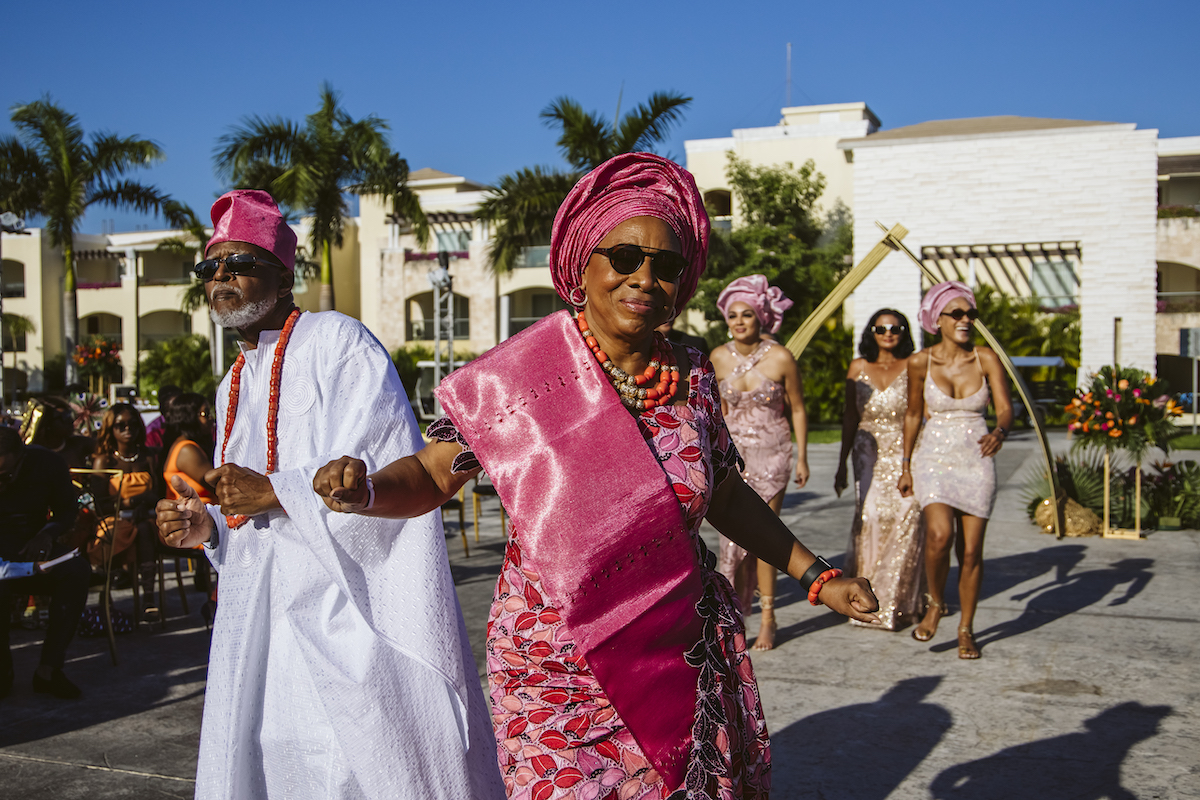 A woman in traditional pink attire and a man in white lead a group of people outdoors, with palm trees and buildings visible in the background.