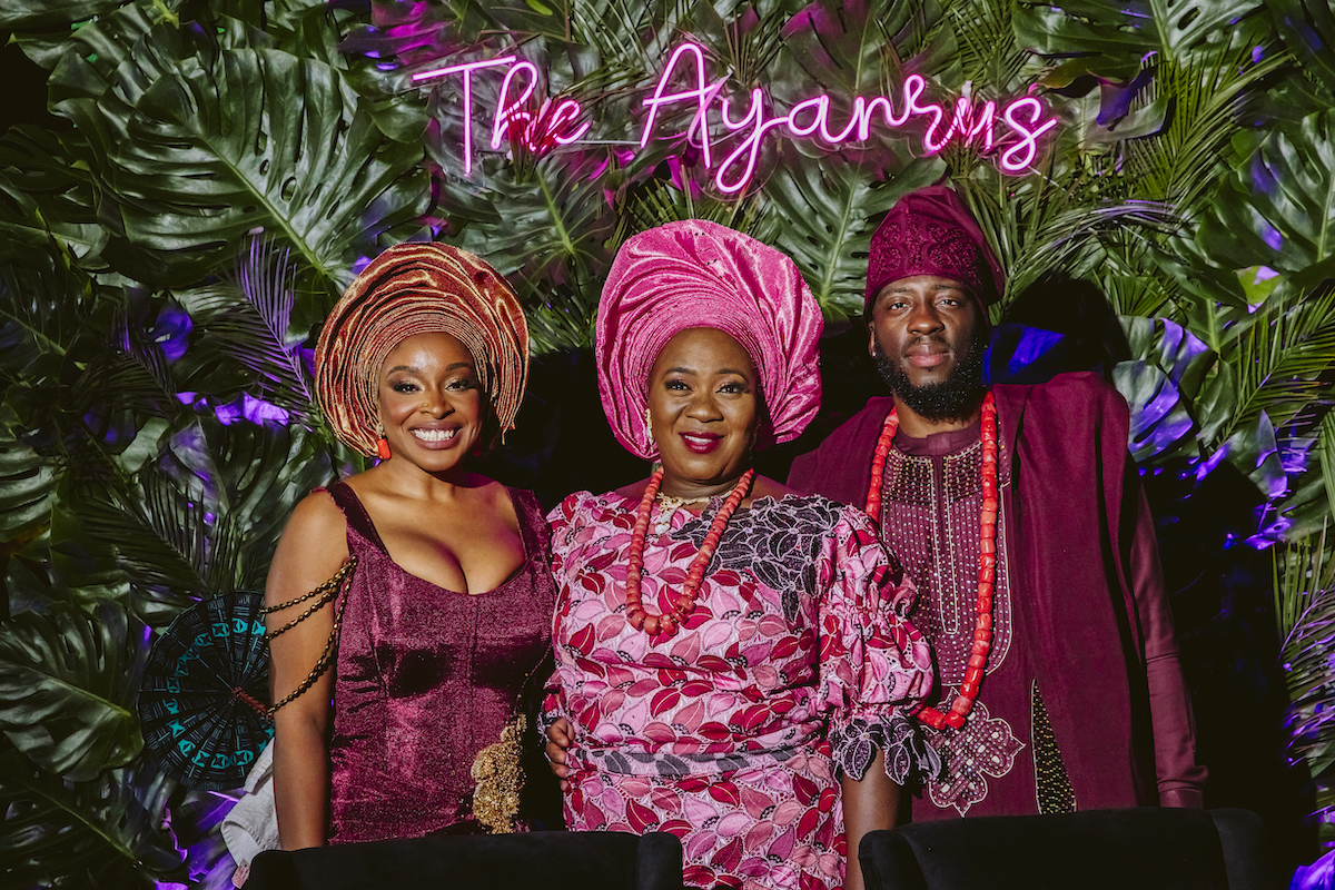 Three people dressed in traditional Nigerian attire stand in front of a leafy backdrop with a neon sign that reads "The Ayanrus."