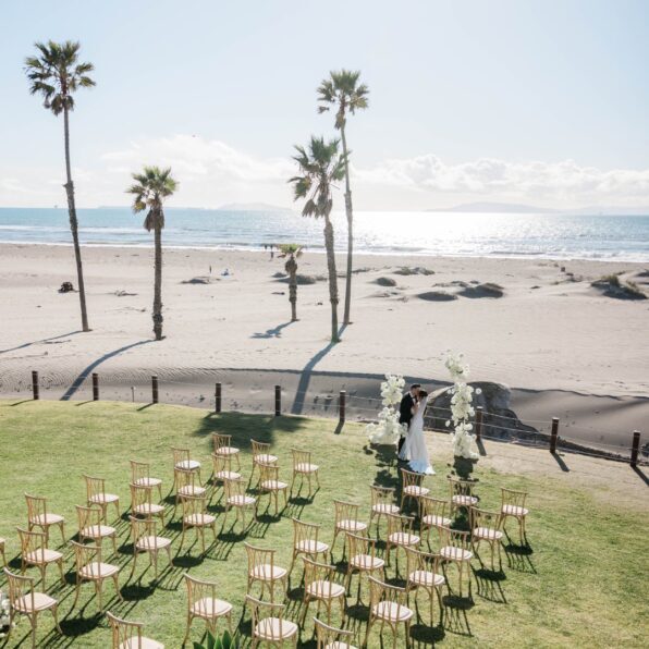 A couple stands under a floral wedding arch on a grassy area near the beach, with empty chairs arranged in rows and palm trees along the shoreline.