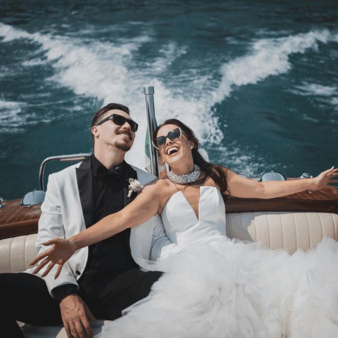 A couple dressed in formal wedding attire smiles and relaxes on a boat, with water and waves visible in the background. This wedding was planned by Bweddings.