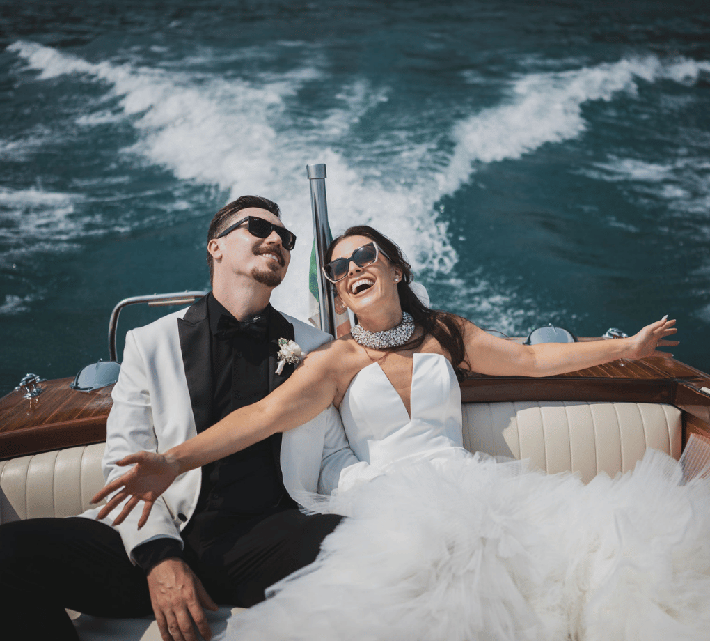 A couple dressed in formal wedding attire smiles and relaxes on a boat, with water and waves visible in the background. This wedding was planned by Bweddings.