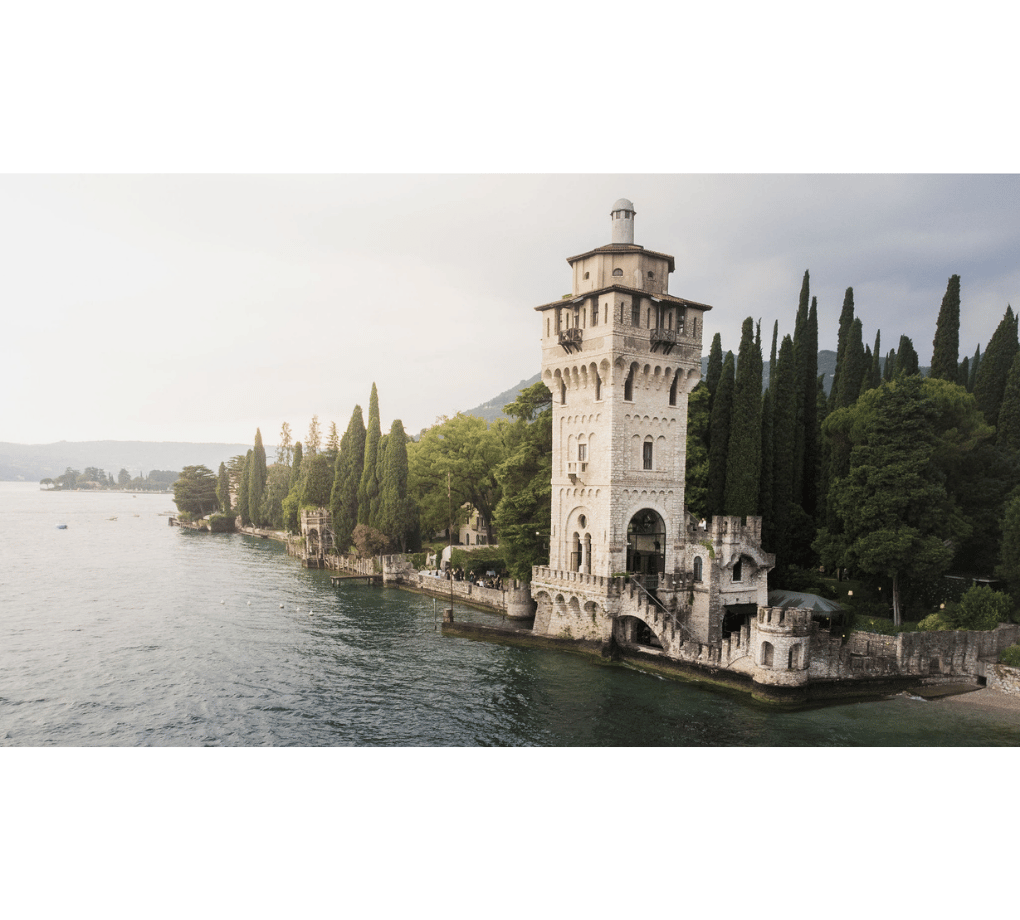 A tall, ornate stone tower stands on the edge of a lake, surrounded by lush trees, with mountains visible in the background.