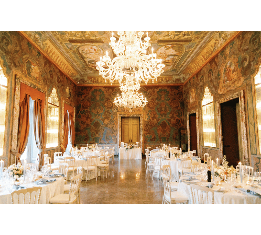 Ornate banquet hall with chandeliers, decorated walls and ceiling, large windows with drapes, and round tables set for a formal event. This wedding was planned by Bweddings.