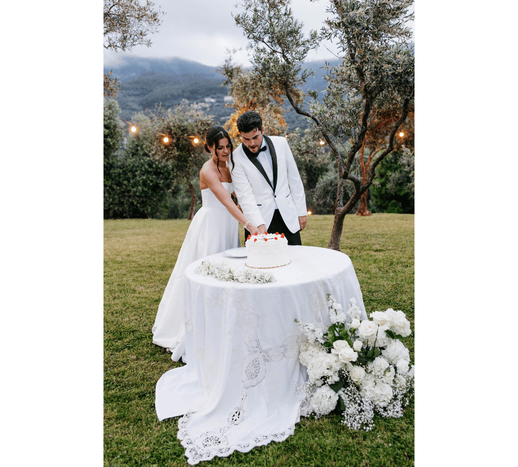 A bride and groom in white formal attire cut a cake together at an outdoor wedding reception, with a floral arrangement and trees in the background. This wedding was planned by Bweddings.